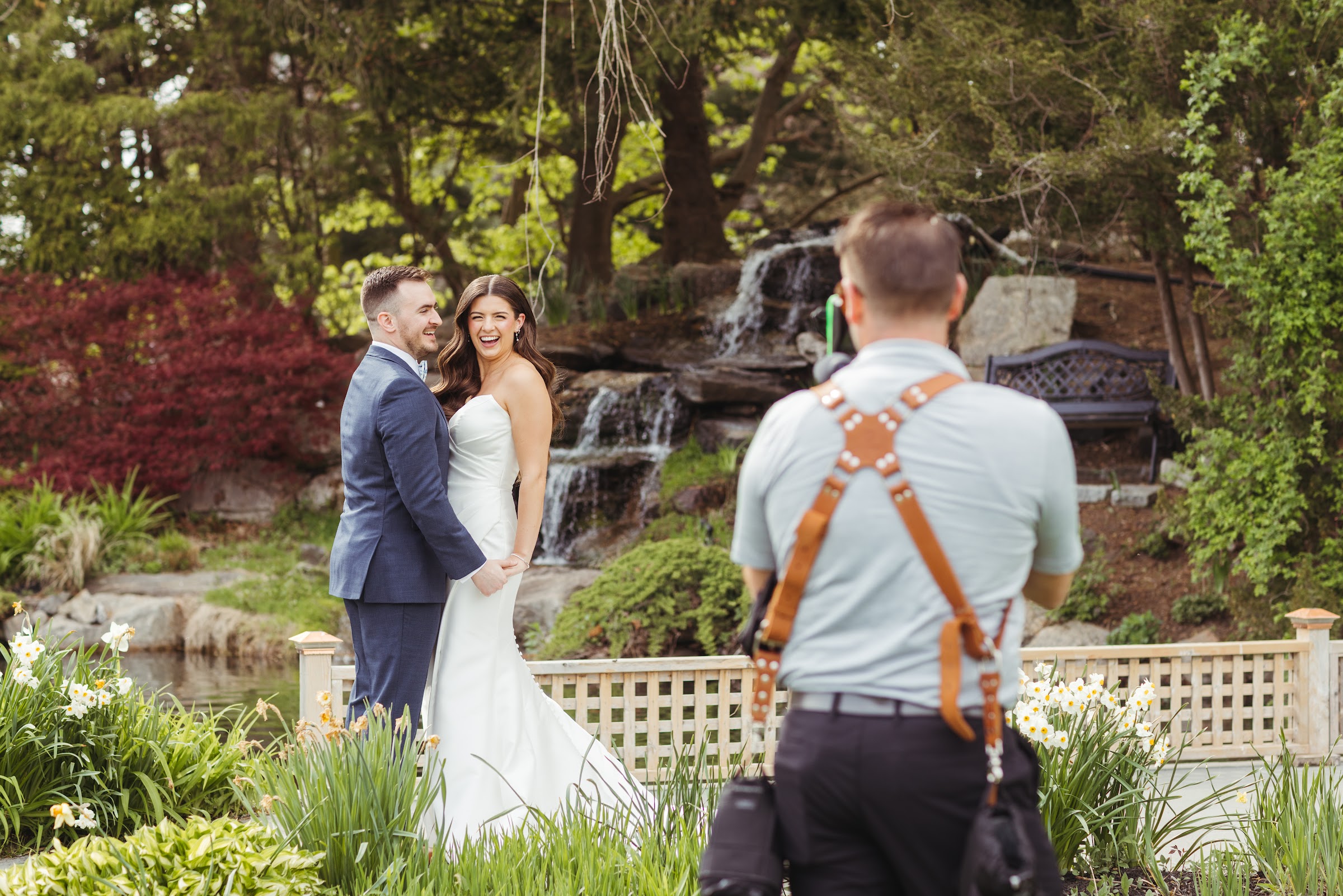 A bride and groom laughing together while being filmed near a garden waterfall.